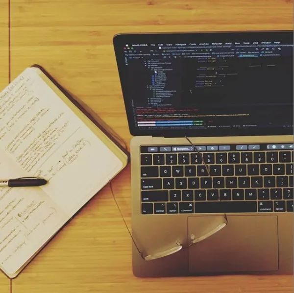 A laptop showing code next to an open notebook on a wooden desk